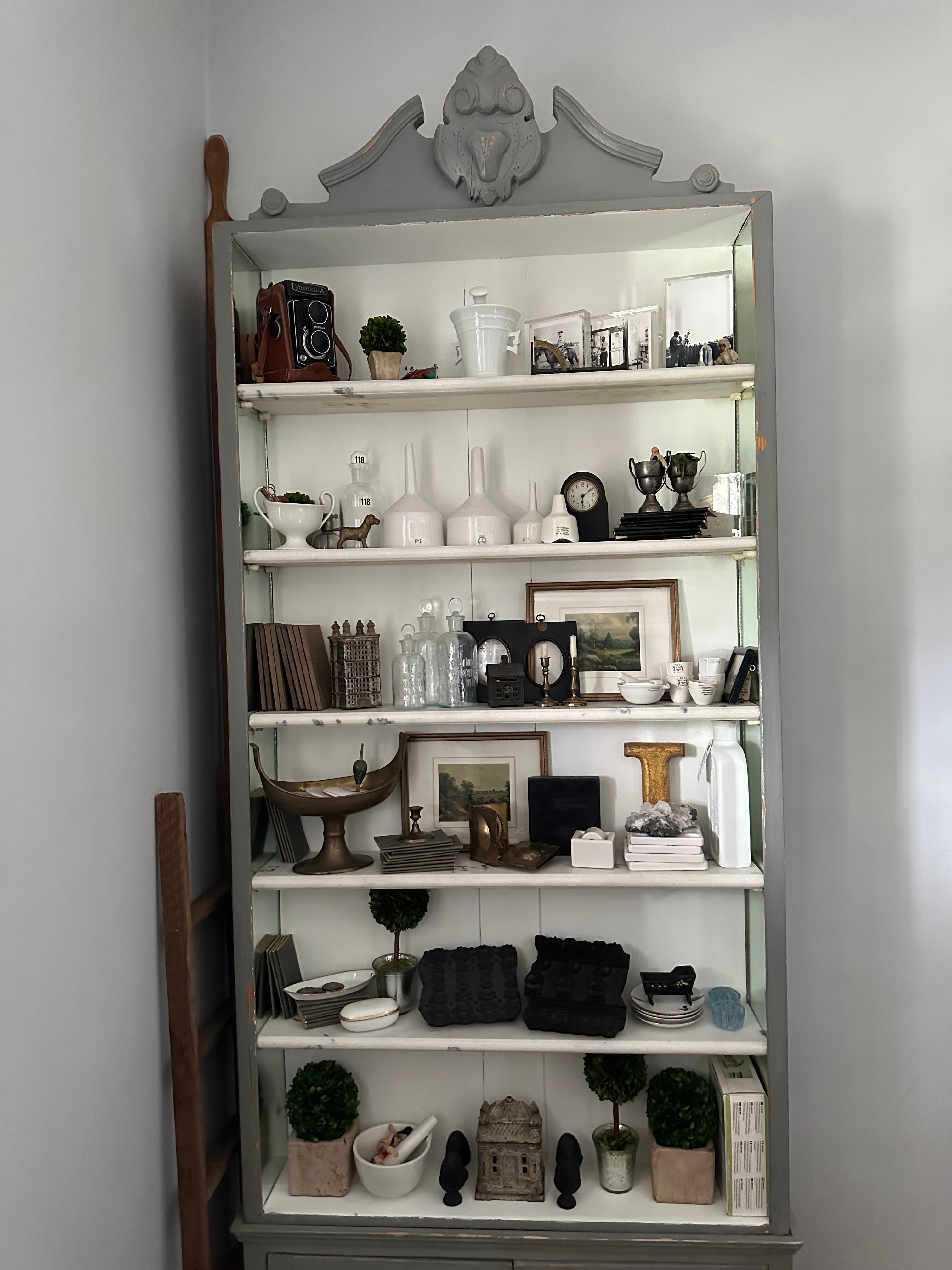 A vintage cabinet filled with decorative items, including framed art, small sculptures, plants, metal cups, vintage cameras, and white ceramics, set against a soft gray wall.