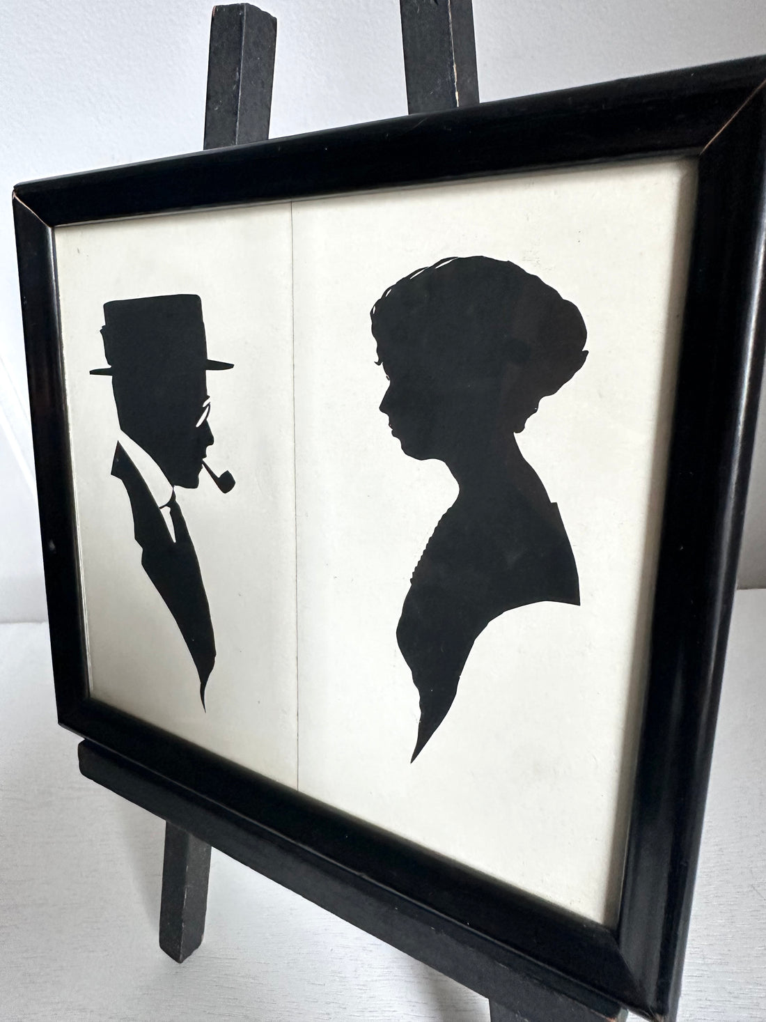 A framed black and white art print featuring silhouette profiles of a man in a top hat and a woman, displayed on a black easel against a light background.