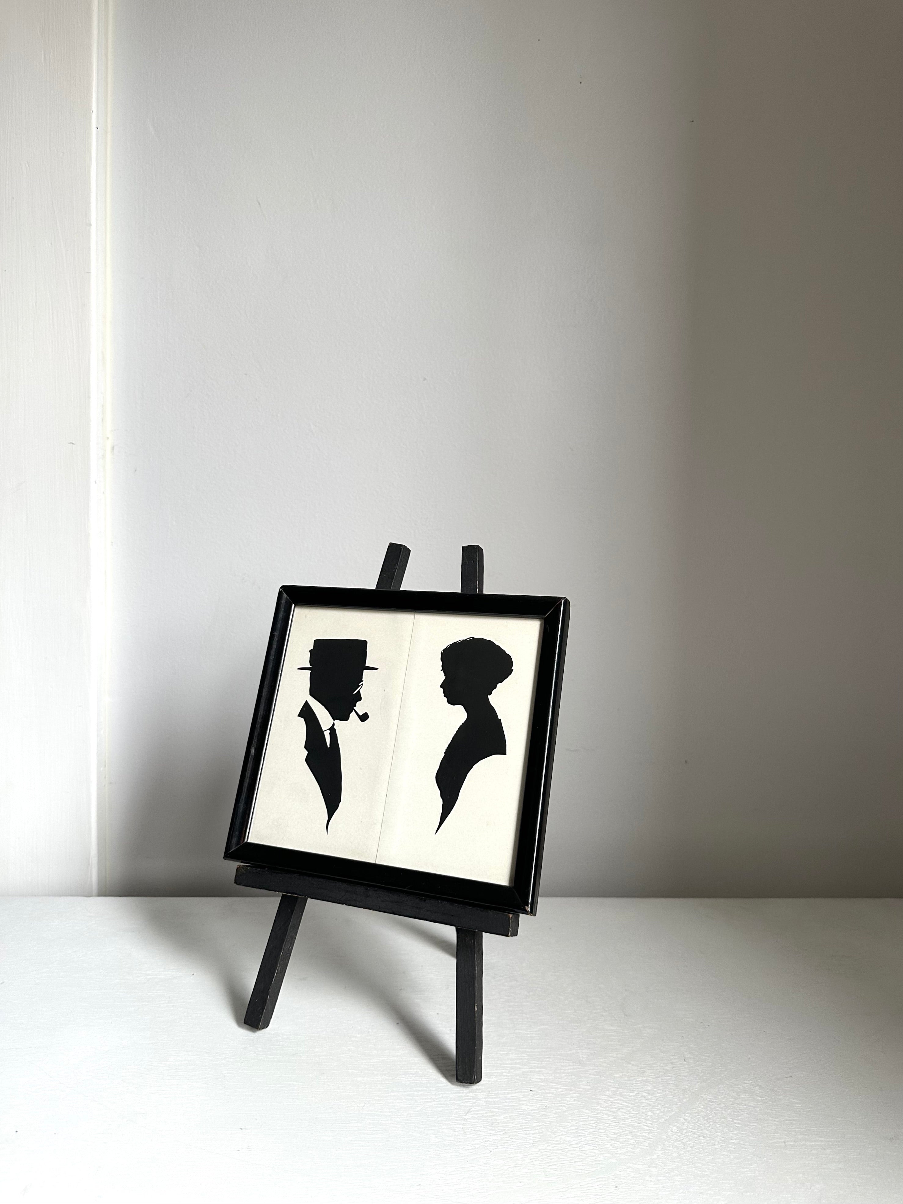 A small black-framed art print displayed on a miniature black easel. The print features black silhouette figures of a man and a woman on a white background.