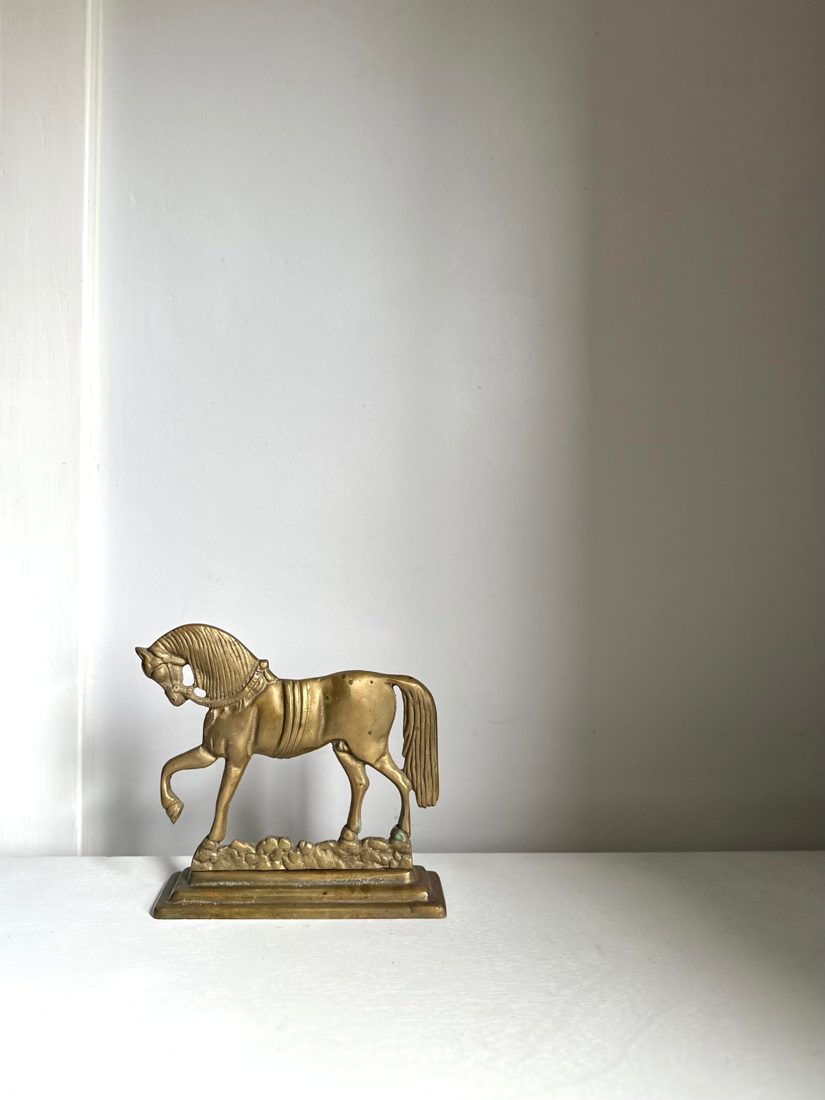 A small metallic horse figurine in a gold finish, standing on a decorative base against a plain light gray wall.