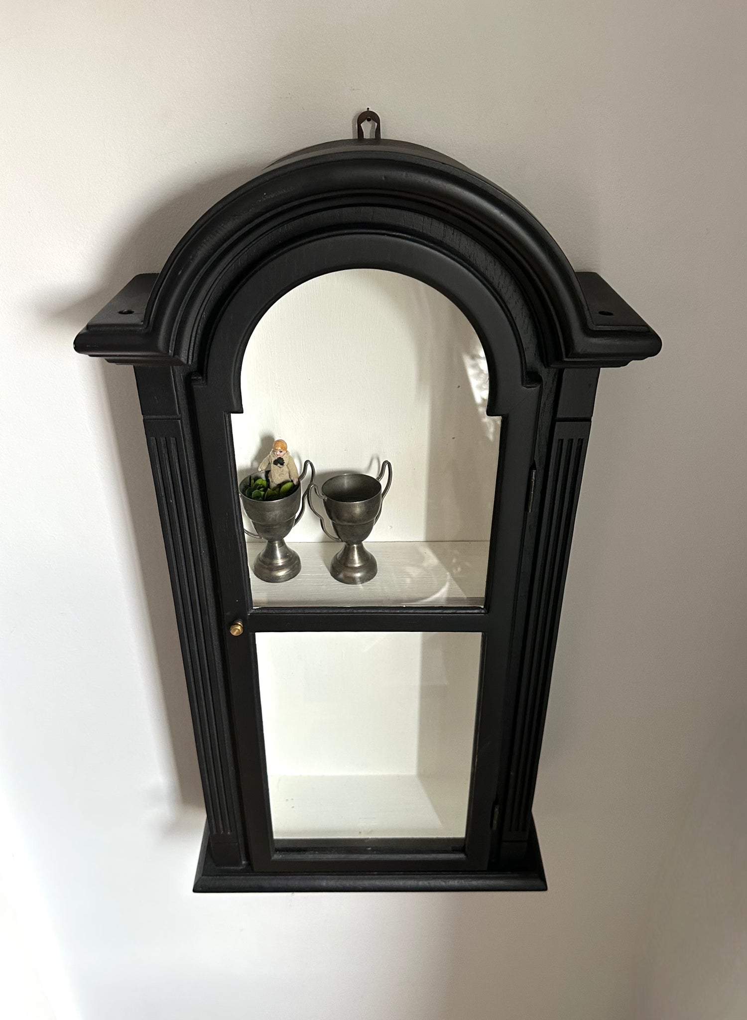 Decorative black arch-shaped display cabinet with door and shelf on a white wall