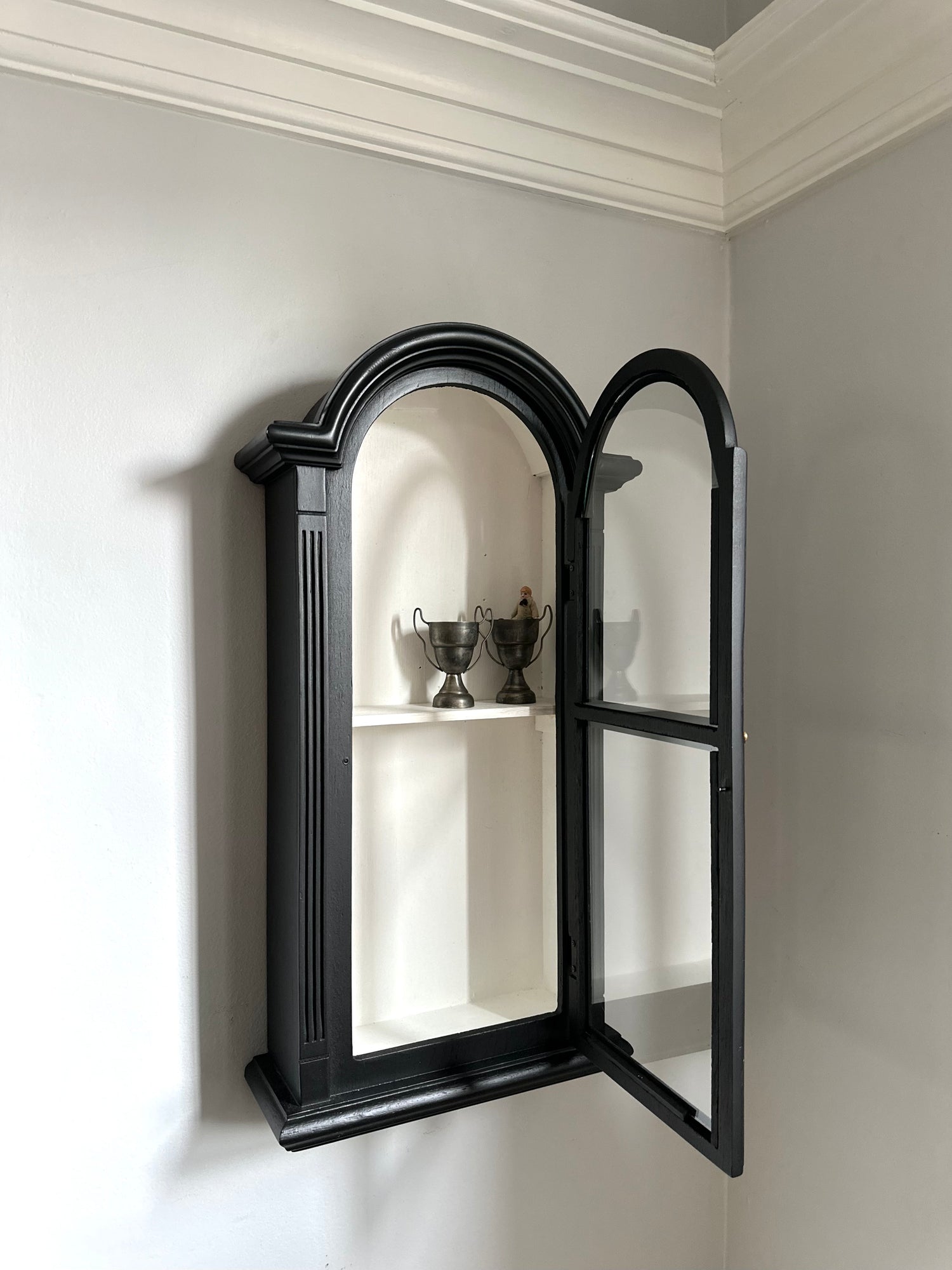 Black wall-mounted cabinet with open doors revealing interior shelves against a light gray wall.