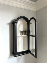 Black wall-mounted cabinet with open doors revealing interior shelves against a light gray wall.