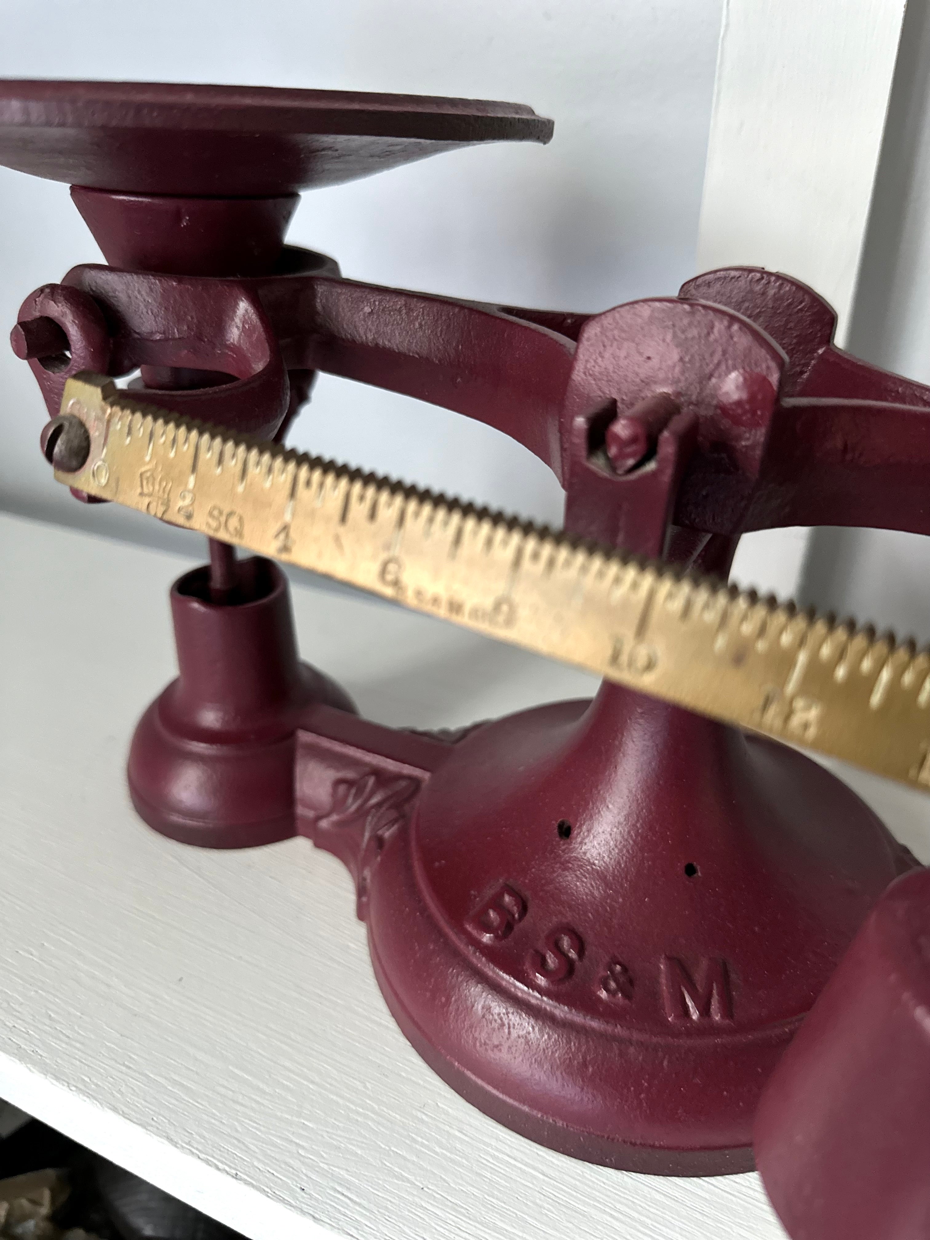 Close-up of an Antique Balance Scale, Made in Hamilton, featuring a red cast iron body with a brass balance beam. It has a round weighing platform and initials B S & M embossed at the base, elegantly displayed on a white shelf.