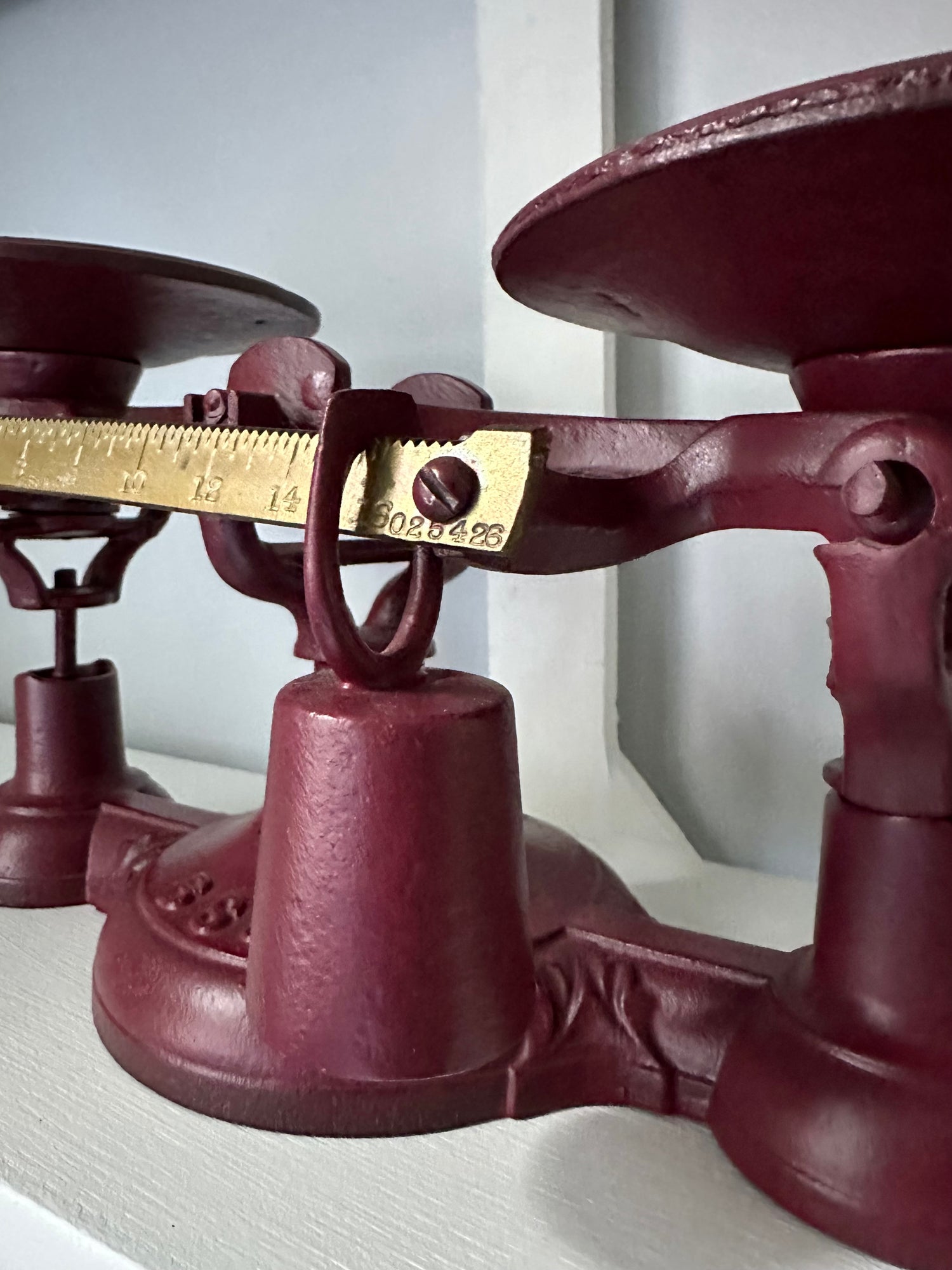 A close-up of an Antique Balance Scale, Made in Hamilton, showcases its red cast iron body with a gold-colored ruler and cylindrical weight. This vintage piece exudes charm as it sits elegantly on a shelf.