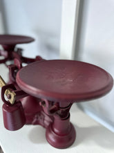 Close-up of the Antique Balance Scale, Made in Hamilton, featuring a vintage red cast iron design with two round metal pans, one in focus. It sits on a white surface, highlighting its detailed mechanical parts and worn texture.