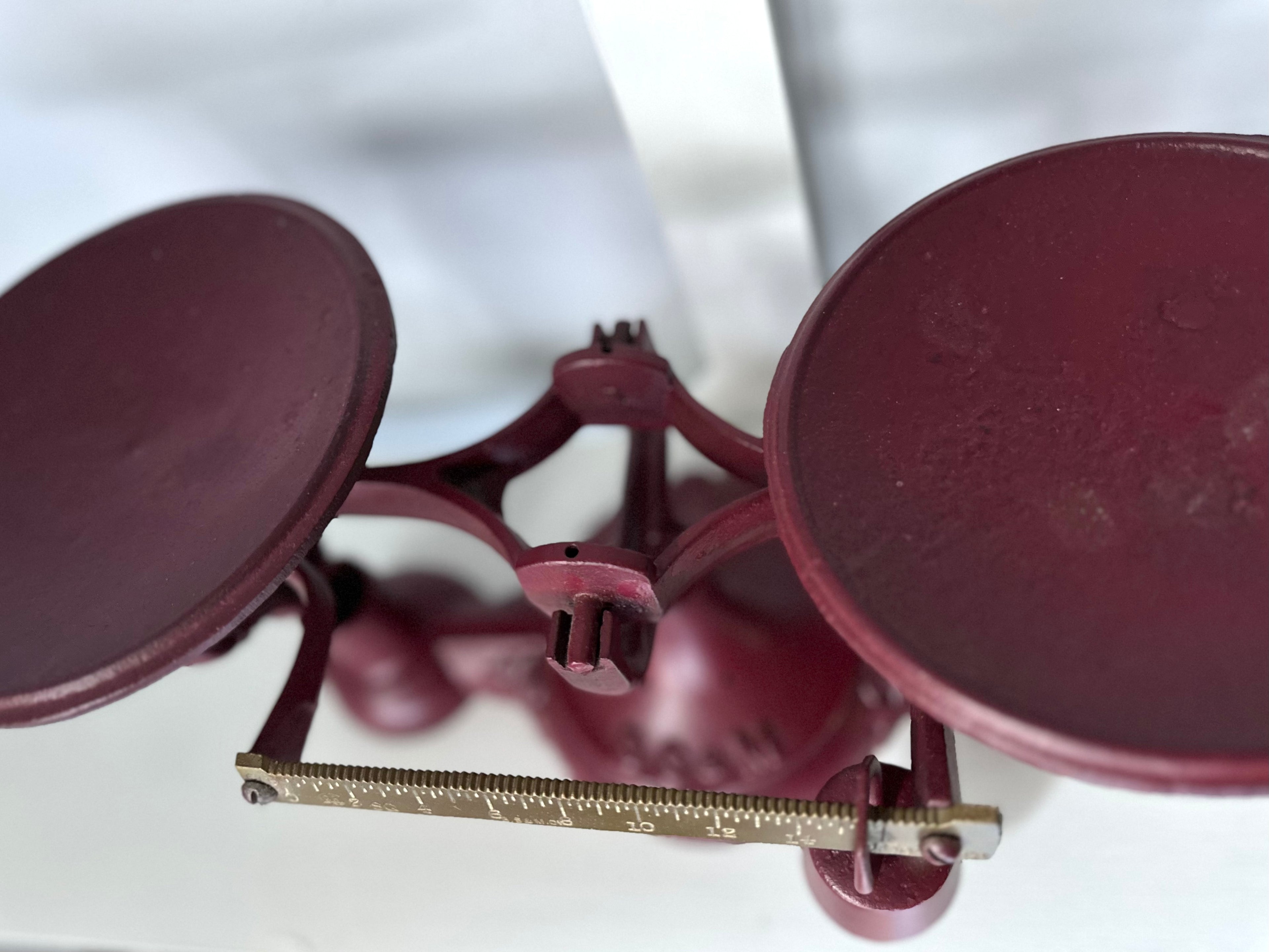 Close-up of an Antique Balance Scale, Made in Hamilton, showcasing its vintage red cast iron design. Featuring two round weighing pans and a brass measuring gauge, it stands out against a white background.