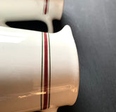 Close-up of the Pair English Stripe Pitchers, featuring ceramic mugs with red and green stripes near the rims, reminiscent of vintage milk pitchers, upside down on a dark surface showcasing their handles as unique kitchen decor.