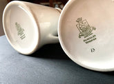 Two white ceramic mugs, styled like vintage milk pitchers called Pair English Stripe Pitchers, sit upside-down on a dark surface. Their undersides display a green logo featuring Hercules Vitrified and a shielded crest, reflecting their classic design and English craftsmanship in kitchen decor.