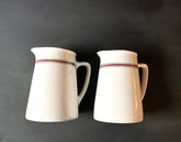 The Pair English Stripe Pitchers are vintage milk pitchers with handles crafted from white ceramic. They each feature a striking red and black stripe near the top, making them ideal for adding classic charm to your kitchen decor against a dark background.