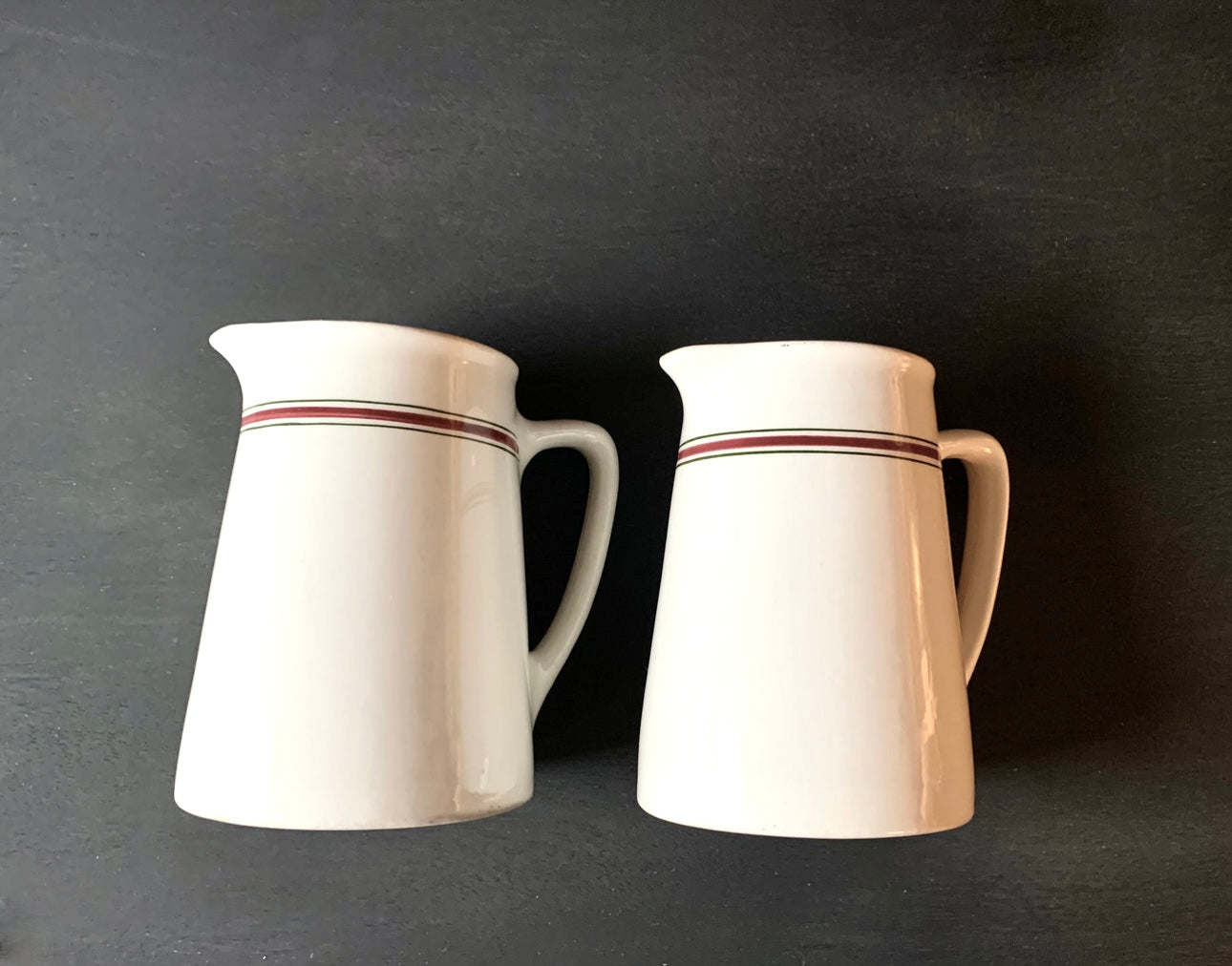 The Pair English Stripe Pitchers are vintage milk pitchers with handles crafted from white ceramic. They each feature a striking red and black stripe near the top, making them ideal for adding classic charm to your kitchen decor against a dark background.