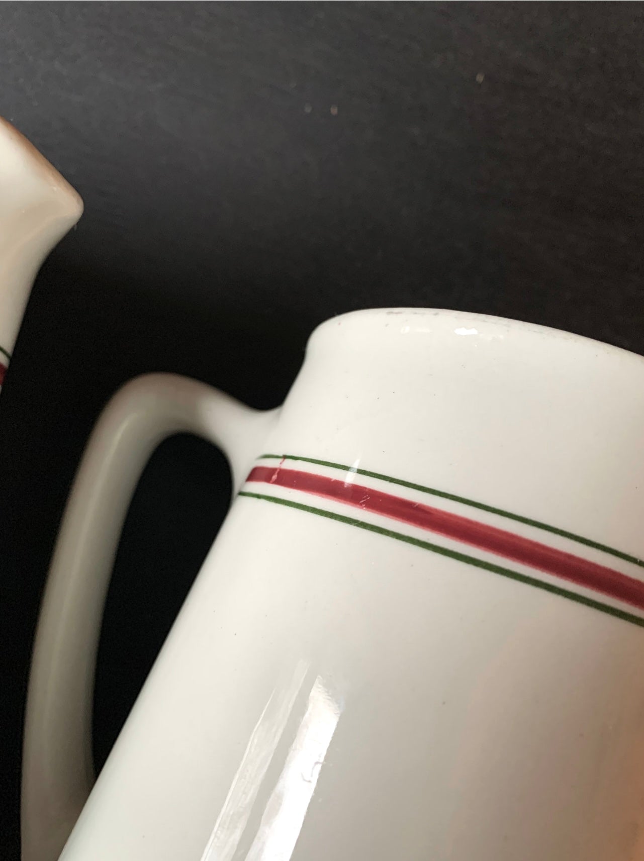 A close-up of a white ceramic pitcher from the Pair English Stripe Pitchers set, against a black background, highlights its simple vintage milk pitcher design with one red and one green stripe near the top. A partially visible handle on the left adds an elegant touch to kitchen decor.