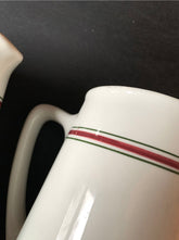 A close-up of a white ceramic pitcher from the Pair English Stripe Pitchers set, against a black background, highlights its simple vintage milk pitcher design with one red and one green stripe near the top. A partially visible handle on the left adds an elegant touch to kitchen decor.