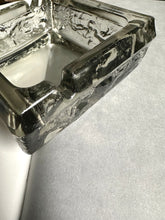 A square transparent green ashtray sitting on an open book with a black and white print in the background