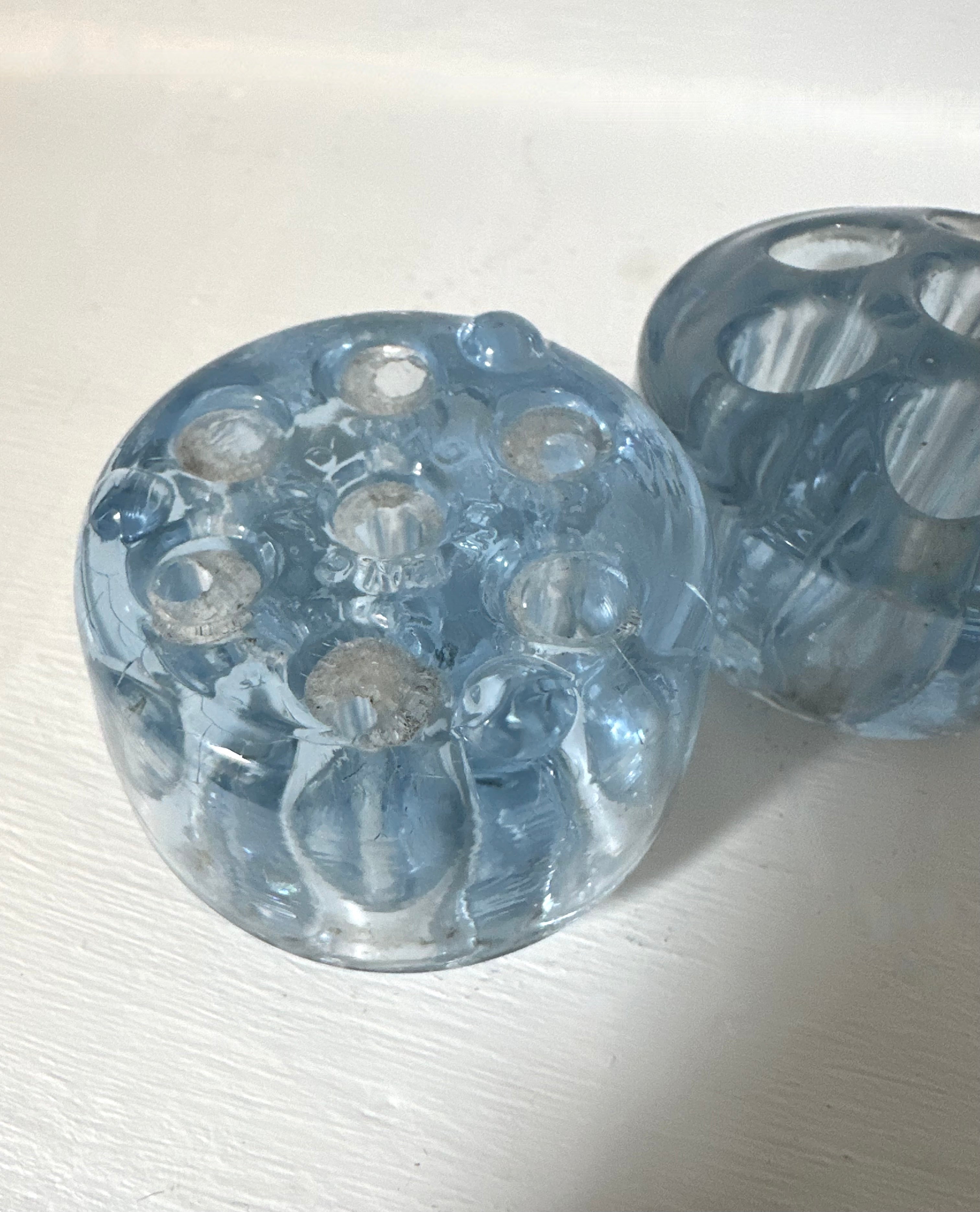 A pair of round, translucent light blue glass candleholders with multiple circular cutouts, displayed on a white textured surface.