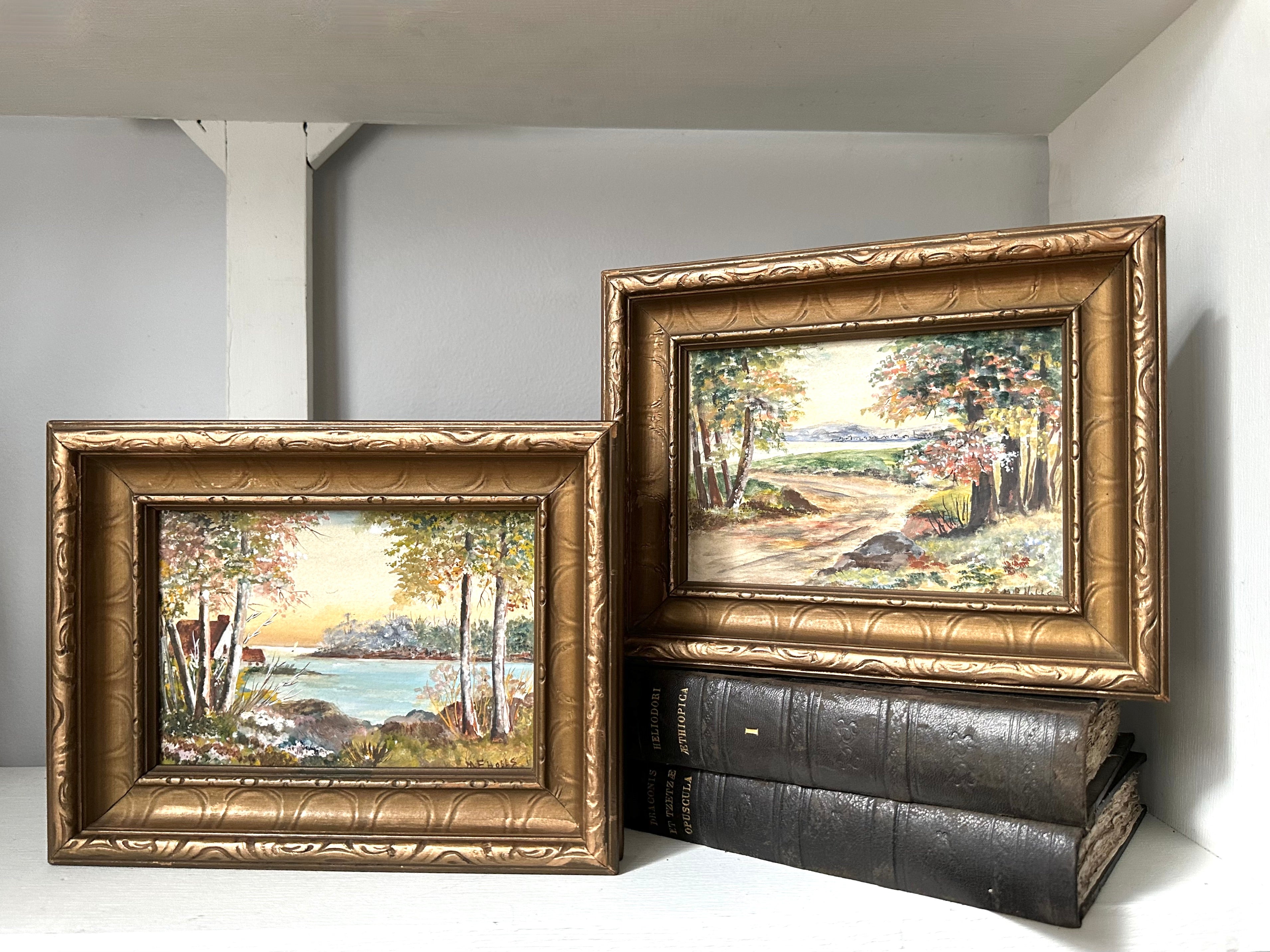 Two framed landscape paintings with ornate gold frames, displayed on a white shelf with dark books underneath the right frame.