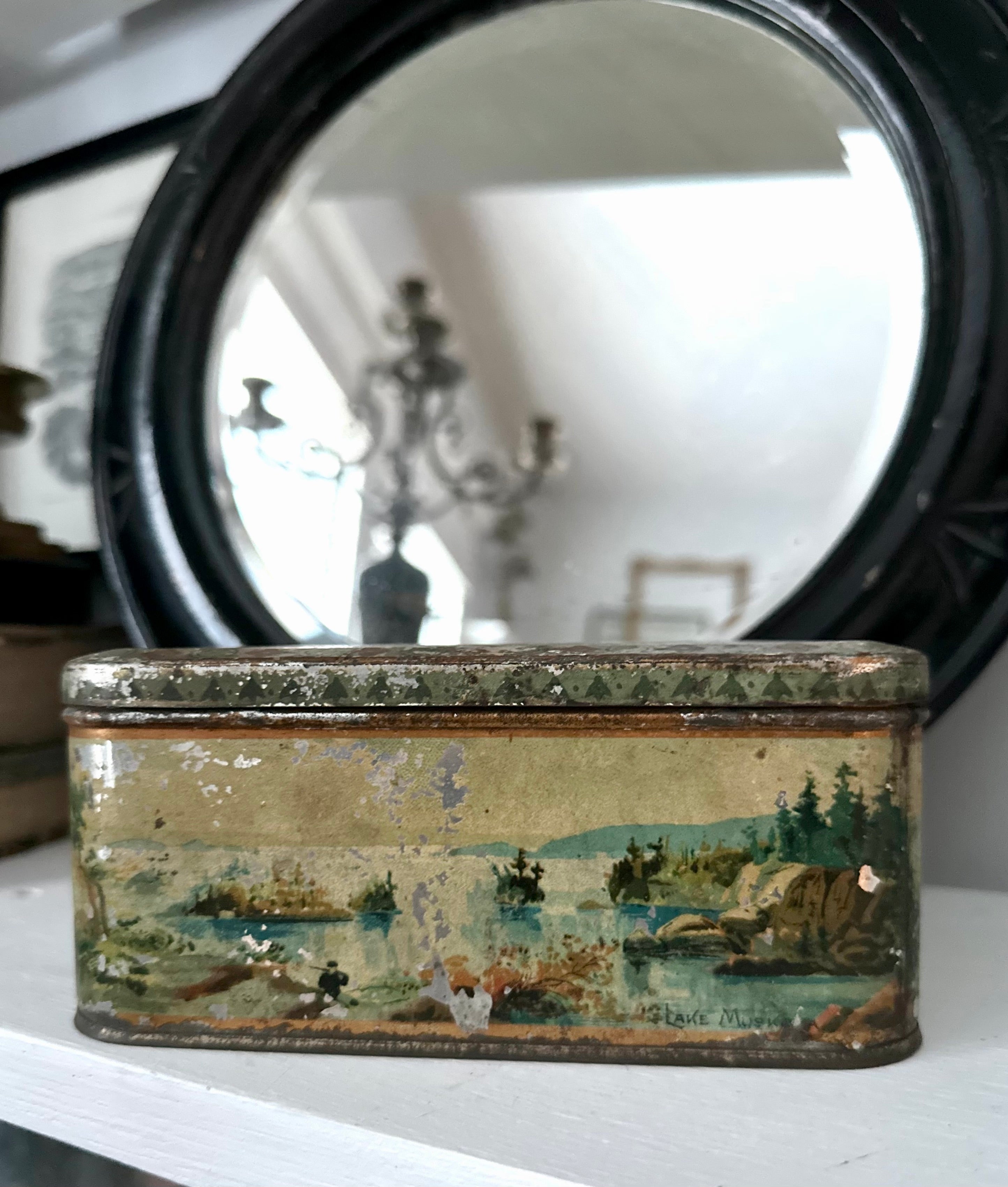 A vintage tin on a white shelf, featuring a scenic landscape design. A round mirror and chandelier are reflected in the background.