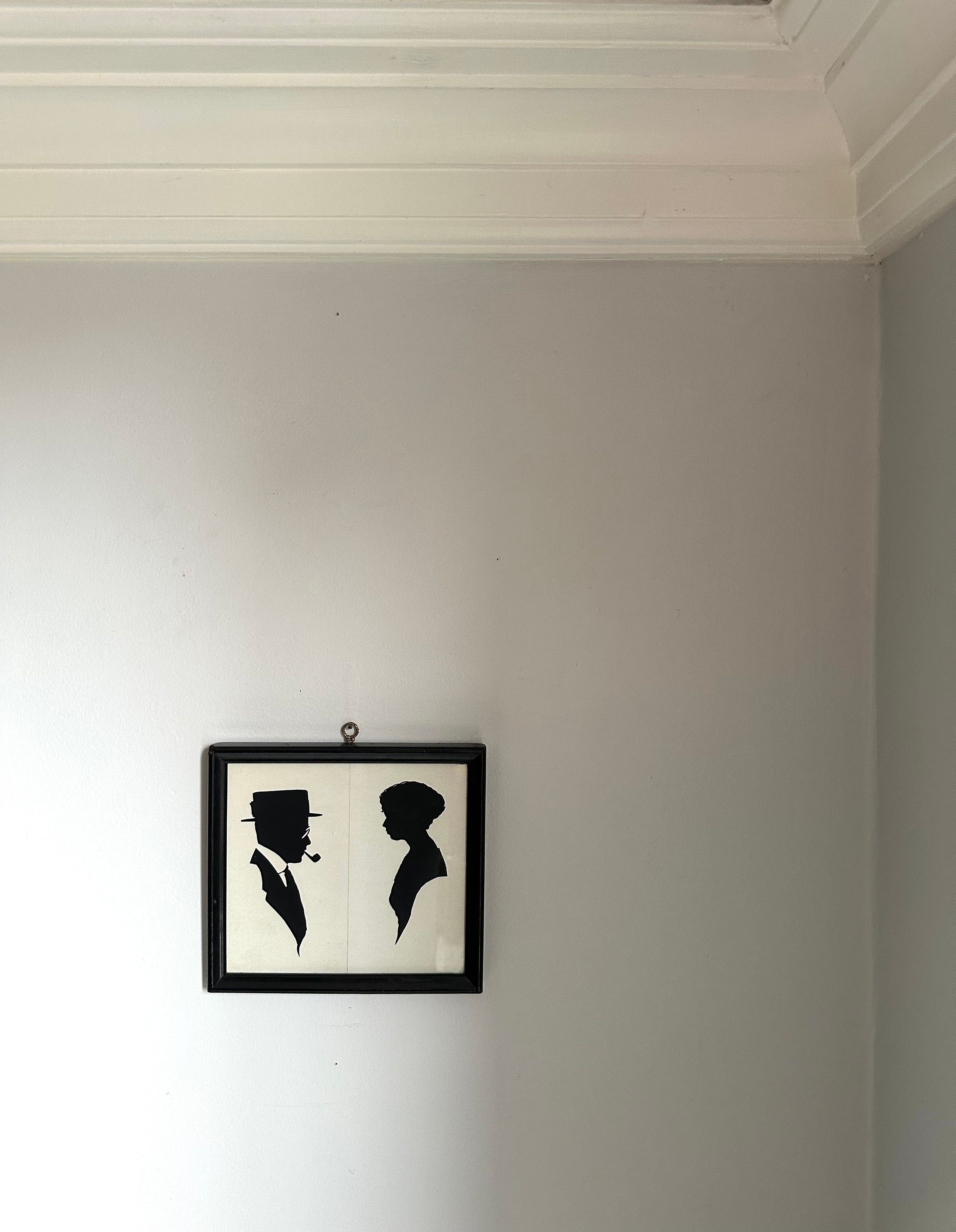 A small black-framed wall art piece hanging on a light gray wall, featuring black silhouette portraits of a man and a woman on a white background.