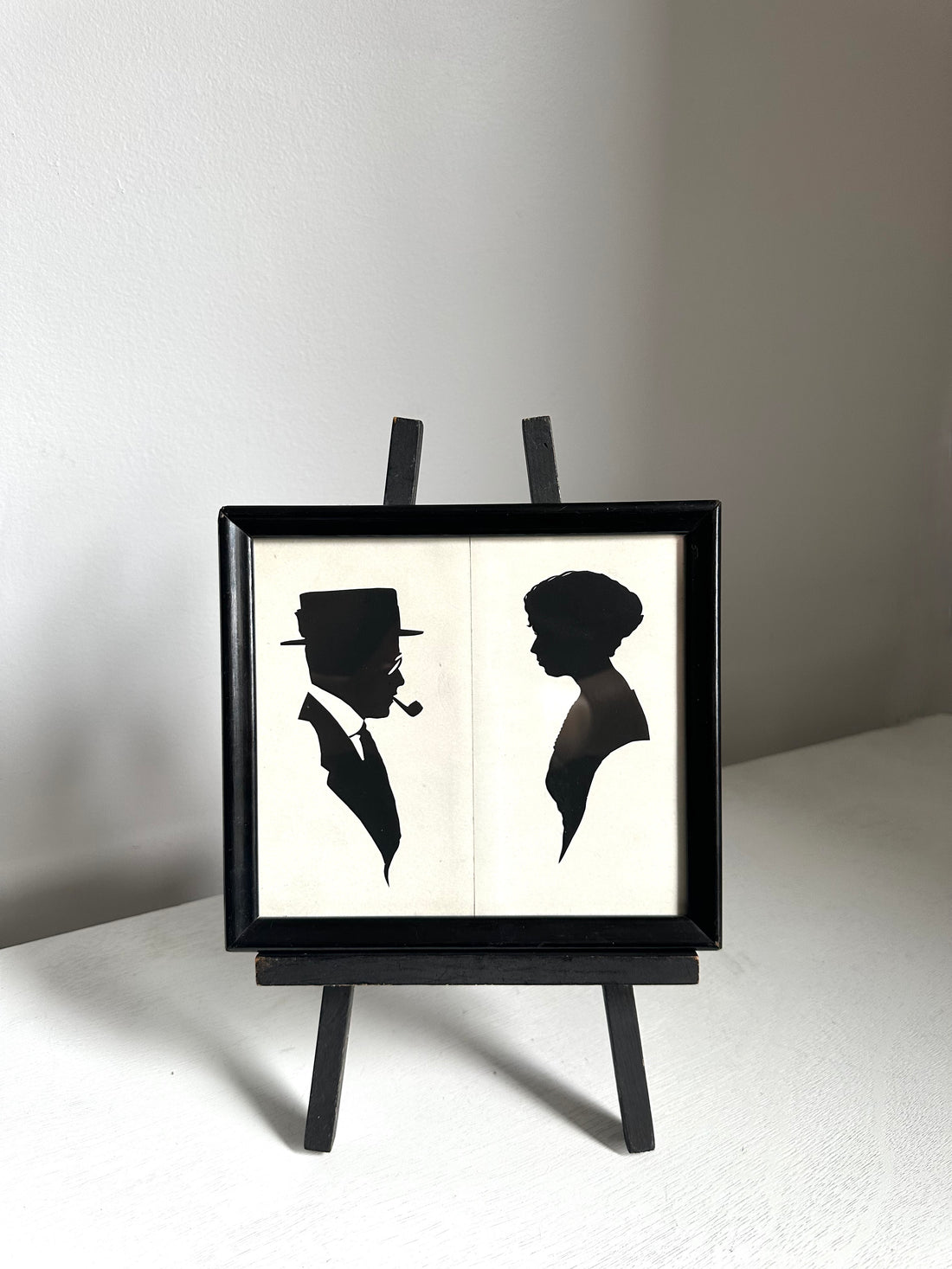 A black-framed wall art piece displayed on a small black easel stand. The artwork shows two black silhouettes on an off-white background: a man in profile wearing a hat and smoking a pipe, and a woman in profile.