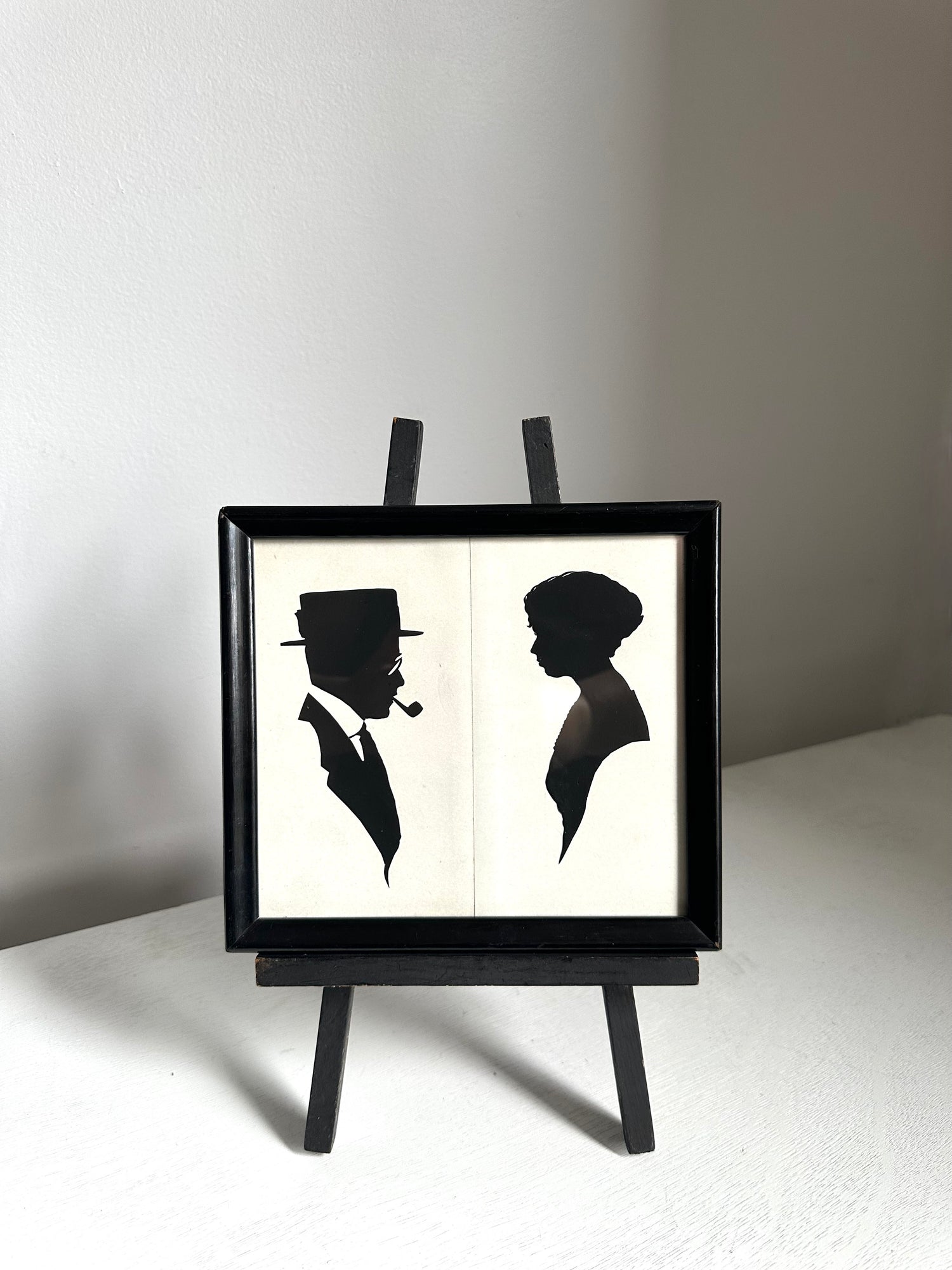 A black-framed wall art piece displayed on a small black easel stand. The artwork shows two black silhouettes on an off-white background: a man in profile wearing a hat and smoking a pipe, and a woman in profile.