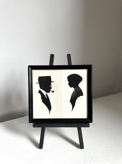 A black-framed wall art piece displayed on a small black easel stand. The artwork shows two black silhouettes on an off-white background: a man in profile wearing a hat and smoking a pipe, and a woman in profile.