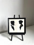 A black-framed wall art piece displayed on a small black easel stand. The artwork shows two black silhouettes on an off-white background: a man in profile wearing a hat and smoking a pipe, and a woman in profile.