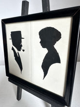 A framed black and white art print featuring silhouette profiles of a man in a top hat and a woman, displayed on a black easel against a light background.