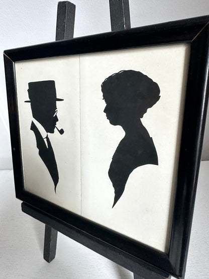 A framed black and white art print featuring silhouette profiles of a man in a top hat and a woman, displayed on a black easel against a light background.