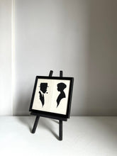 A small black-framed art print displayed on a miniature black easel. The print features black silhouette figures of a man and a woman on a white background.