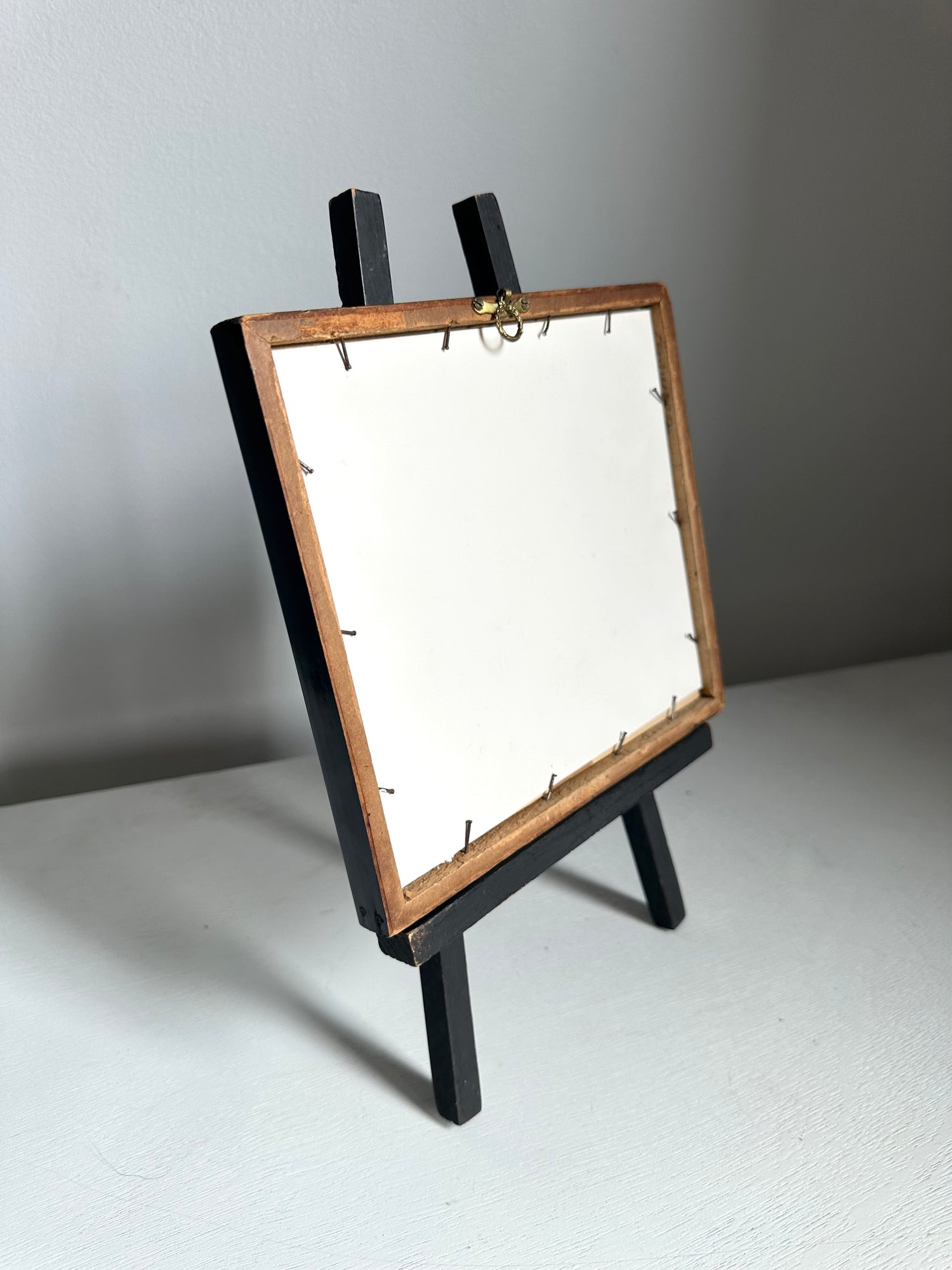 A small tabletop easel stand with a rectangular wooden frame and black legs, shown from the back on a light gray surface against a gray wall.