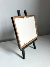 A small tabletop easel stand with a rectangular wooden frame and black legs, shown from the back on a light gray surface against a gray wall.