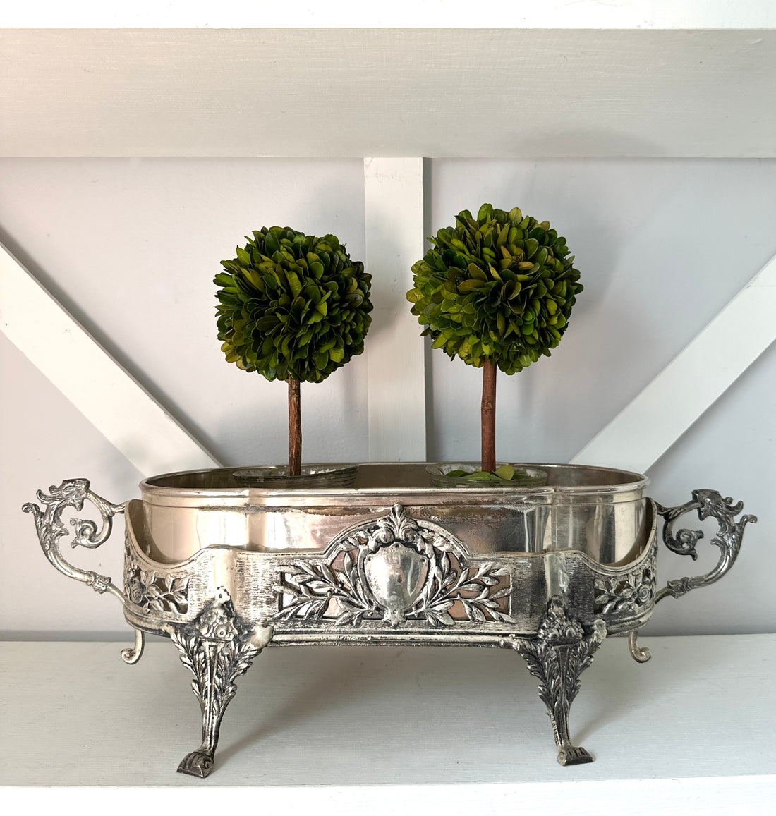 An ornate silver metal planter with decorative legs and side handles, holding two small green topiary trees, displayed on a white shelf against a white wall.