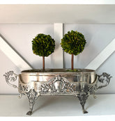 An ornate silver metal planter with decorative legs and side handles, holding two small green topiary trees, displayed on a white shelf against a white wall.