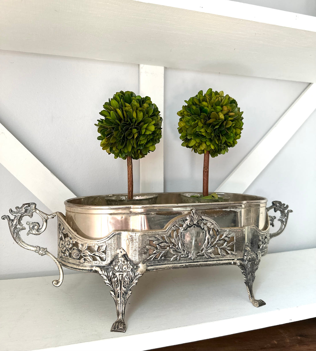 An ornate silver metal planter with carved floral details and two small green topiary trees in it, displayed on a white shelf against a white wall.