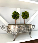 An ornate silver metal planter with carved floral details and two small green topiary trees in it, displayed on a white shelf against a white wall.