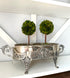 An ornate silver metal planter with carved floral details and two small green topiary trees in it, displayed on a white shelf against a white wall.