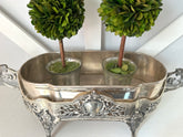 An ornate silver metal tray with decorative handles and legs, holding two small potted green topiary trees against a white paneled background.