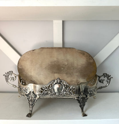 An ornate vintage silver pedestal stand with a round beige stone or ceramic top, featuring decorative scrollwork sides and four claw feet, displayed on a white shelf.