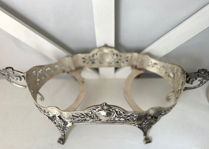 An ornate silver metal stand with decorative filigree cutouts, scrollwork, and small feet, photographed against a white background.
