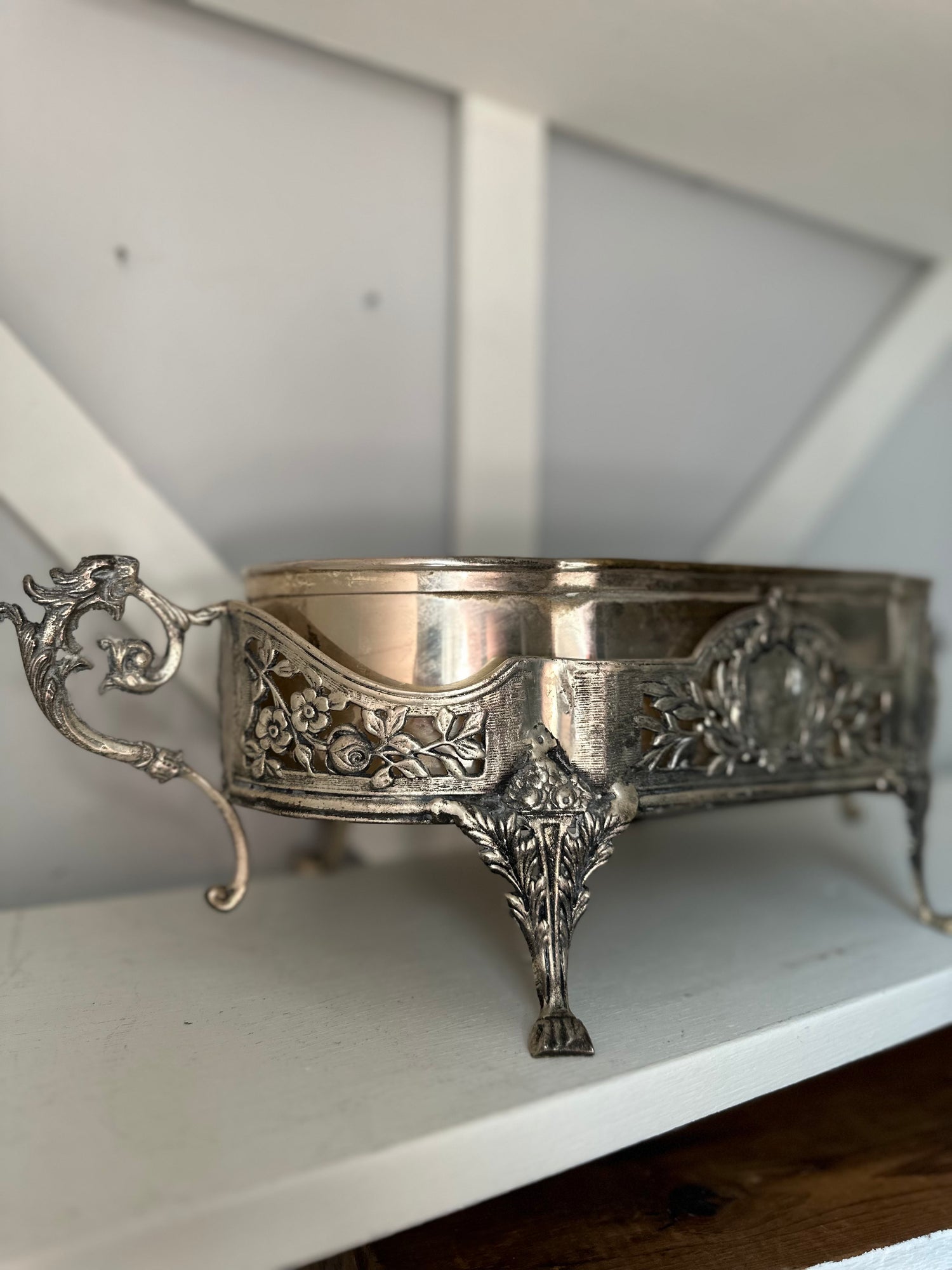An ornate silver-toned metal bowl with decorative floral relief patterns and a curved handle, displayed on a white shelf.