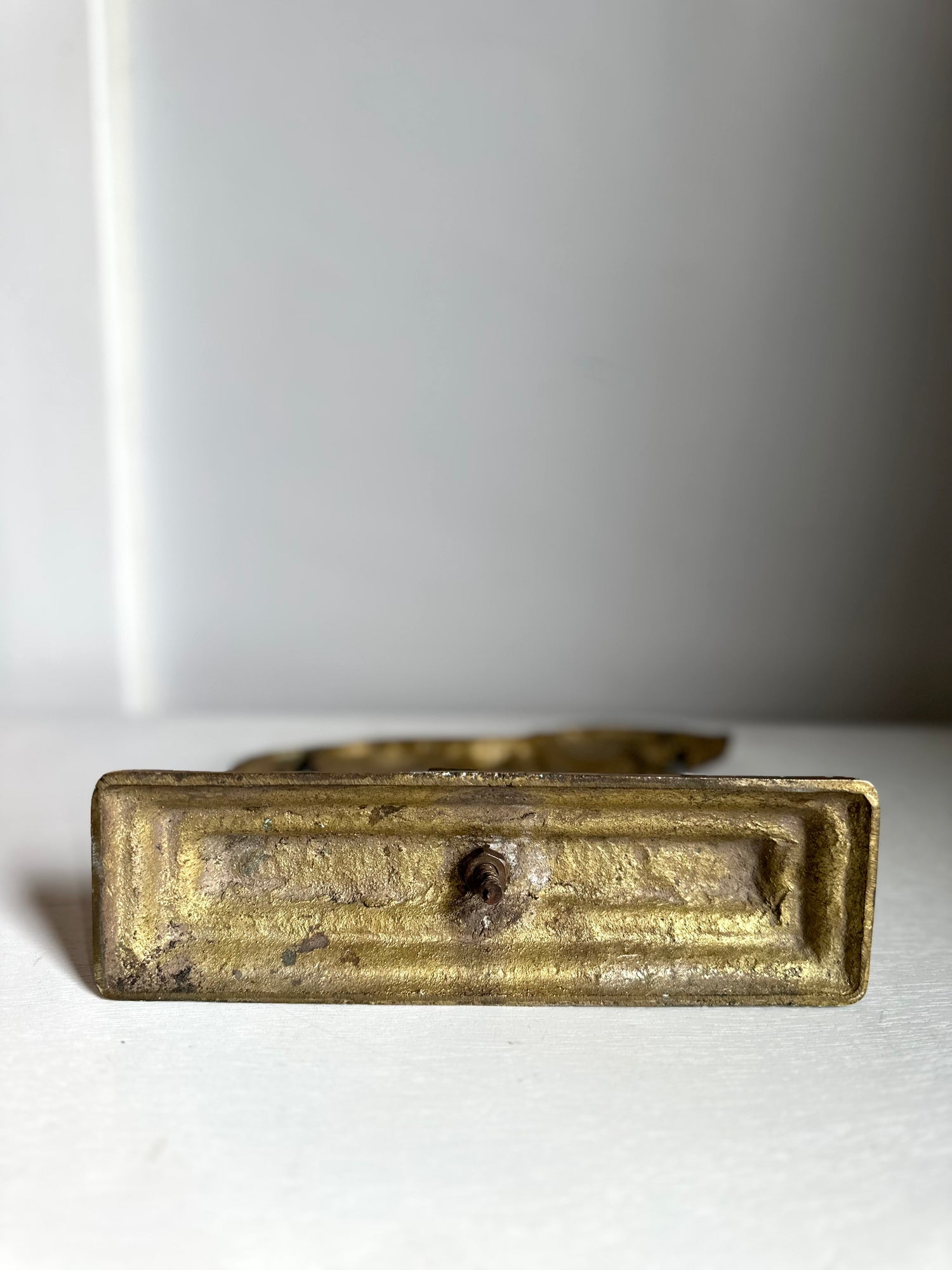 An antique brass drawer pull handle with a rectangular plate and a central round knob, resting on a white surface against a light gray background.