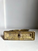 An antique brass drawer pull handle with a rectangular plate and a central round knob, resting on a white surface against a light gray background.