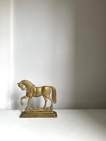 A small metallic horse figurine in a gold finish, standing on a decorative base against a plain light gray wall.