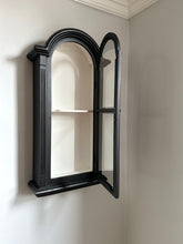 Black-framed antique display cabinet leaning against a light gray wall 