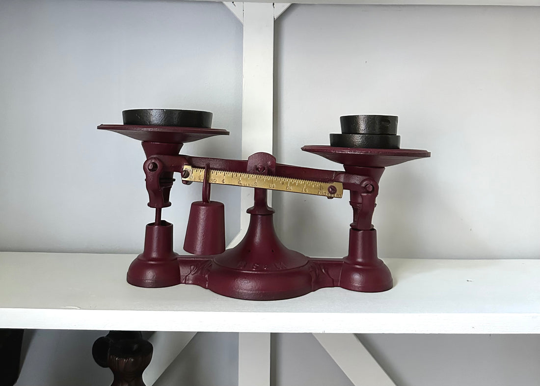 The Antique Balance Scale, made in Hamilton, sits on a white shelf. It features a vintage red cast iron design with two round black weights and a brass beam decorated with precise measurement markings.