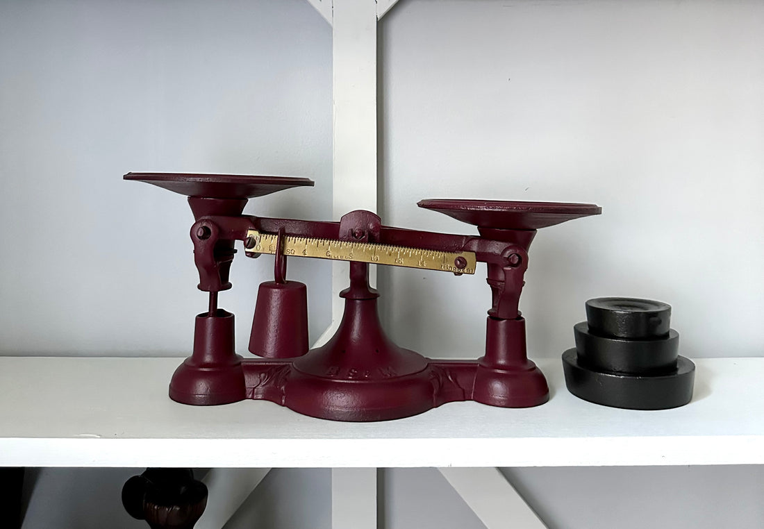 Displayed on a white shelf is an Antique Balance Scale, Made in Hamilton. Crafted from red cast iron, it features two round pans and a brass measuring beam, accompanied by several black circular weights.