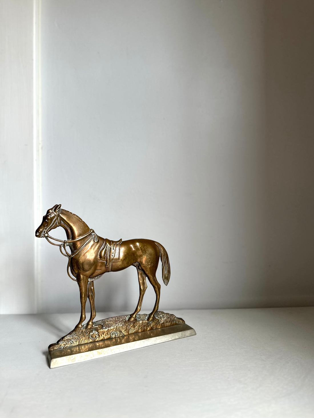 A small bronze horse figurine standing on a textured base, photographed against a plain light gray wall and surface.