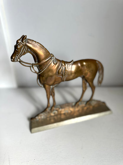 A small metallic horse figurine or sculpture in a warm bronze-gold finish, standing on a textured base against a plain light background.