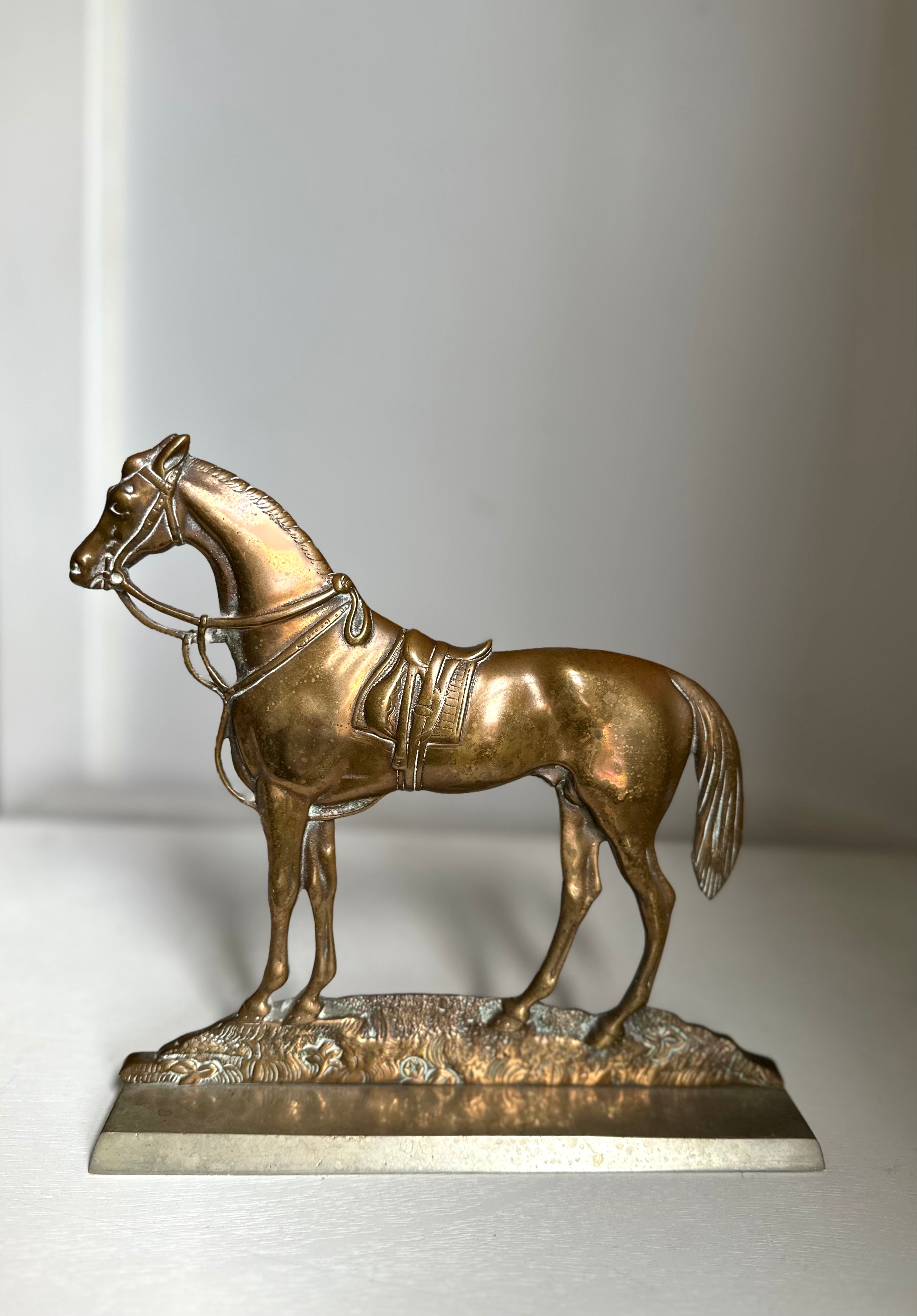 A metallic horse figurine sculpture in a bronze finish, standing on a textured base with a polished rectangular pedestal, photographed against a neutral light gray background.