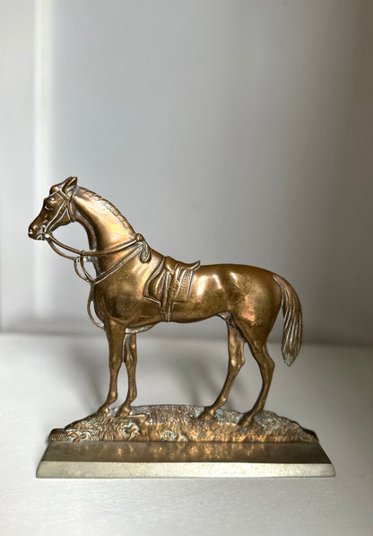 A metallic horse figurine sculpture in a bronze finish, standing on a textured base with a polished rectangular pedestal, photographed against a neutral light gray background.